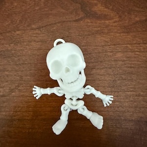 May include: A white, articulated skeleton figurine featuring a skull with a wide, toothy grin, jointed arms, and jointed legs. The figure includes a small loop at the top of the cranium, allowing it to function as a hanging ornament or charm.