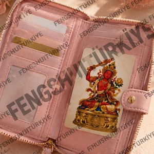May include: A light pink bi-fold wallet featuring a gold-tone snap closure, multiple interior card slots, and a zippered compartment. A rectangular card depicting a red deity figure in a seated position is tucked into the clear ID window slot.