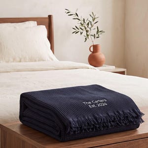 May include: A folded navy blue waffle-weave blanket with fringe edges. The blanket features white embroidered text that reads The Carters Est. 2024. The fabric has a distinct grid-like texture throughout the surface of the textile material.