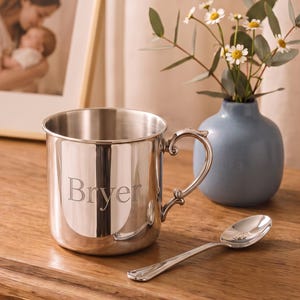 May include: A polished silver-toned metal mug featuring the engraved name Bryer on the front, accompanied by a matching silver-toned spoon. The mug has a smooth, cylindrical body and a decorative, curved handle attached to the side.