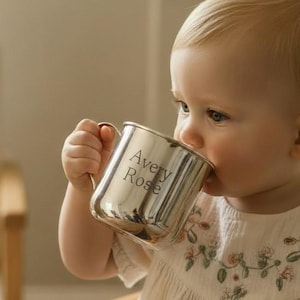Peut inclure: Une tasse pour bébé en métal argenté avec le nom "Avery Rose" gravé sur le côté. La tasse est tenue par un bébé portant une robe blanche avec des broderies florales. Le bébé est assis dans une chaise haute en bois.