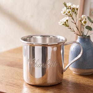 May include: A polished silver-toned metal mug featuring a single handle and a smooth, reflective surface. The name Charlotte is engraved in a cursive script on the front of the mug. The mug has a cylindrical shape with a slightly flared rim.
