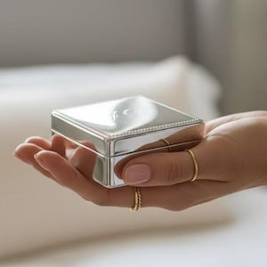 Könnte beinhalten: Eine kleine, quadratische Box aus poliertem, silberfarbenem Metall mit einem Klappdeckel und einer perlenartigen Verzierung entlang der Oberkante. Die Box hat eine reflektierende, spiegelnde Oberfläche und eine klare, geometrische Form.
