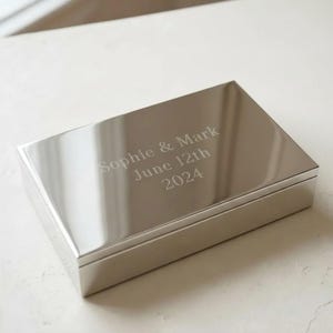 May include: A rectangular silver-toned metal box with a polished, reflective surface. The lid is engraved with the text Sophie & Mark, June 12th, 2024. The box features clean, sharp edges and a smooth finish, presenting a simple and functional design.