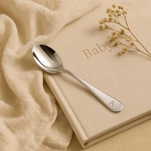 May include: A polished silver spoon with a rounded bowl and a slender handle, lying on a beige book embossed with the word "Baby". The handle is engraved with the letter "A". A sprig of dried flowers is placed on the book.