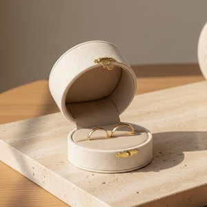 Op de afbeelding: Een ronde, beige met stof beklede juwelendoos met een open deksel, voorzien van een goudkleurige metalen sluiting en scharnier. De binnenkant is bekleed met bijpassend beige fluweel en bevat twee gleuven met twee eenvoudige, gouden ringen.
