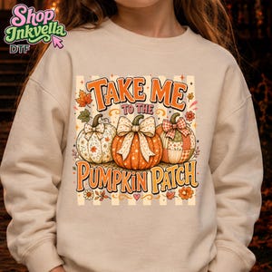 May include: A beige crewneck sweatshirt featuring a graphic print of three pumpkins with bows, surrounded by autumn leaves and flowers. The text reads "TAKE ME TO THE PUMPKIN PATCH" in orange letters. The background of the graphic has vertical stripes.