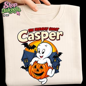 Könnte beinhalten: Ein cremefarbenes T-Shirt mit einer Grafik von Casper dem freundlichen Geist, der in einem geschnitzten orangefarbenen Kürbis sitzt. Das Design zeigt ein dunkelblaues Spukhaus, schwarze Fledermäuse, einen gelben Mond und den Text THE FRIENDLY GHOST Casper.