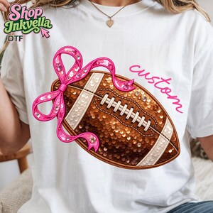 May include: A white t-shirt featuring a graphic of a brown sequined football adorned with a bright pink sequined bow. The football has white sequined laces and stripes. The word custom is written in pink cursive script to the right of the football.