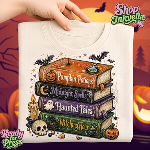 May include: A cream-colored t-shirt featuring a graphic of four stacked books with the titles Pumpkin Potions, Midnight Spells, Haunted Tales, and Witching Hour. The design includes a candle, a skull, bats, spiders, and small pumpkins.