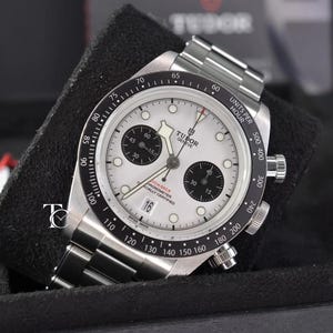 Könnte beinhalten: Eine Tudor Geneve Chronographen-Armbanduhr mit weißem Zifferblatt, zwei schwarzen Hilfszifferblättern und einer schwarzen Tachymeterlünette. Die Uhr verfügt über ein Edelstahlgehäuse und -armband, ein Datumsfenster bei sechs Uhr und Leuchtindizes.