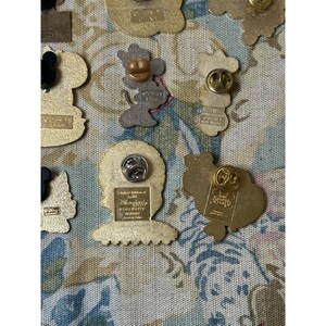 May include: A collection of Disney pins with various character designs. The pins are gold-toned and feature images of Mickey Mouse, Minnie Mouse, and other characters. Some pins have text, and all have pin backs.