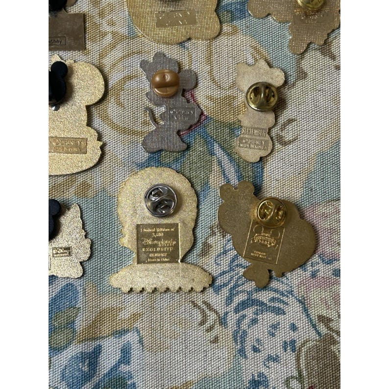 May include: A collection of Disney pins with various character designs. The pins are gold-toned and feature images of Mickey Mouse, Minnie Mouse, and other characters. Some pins have text, and all have pin backs.