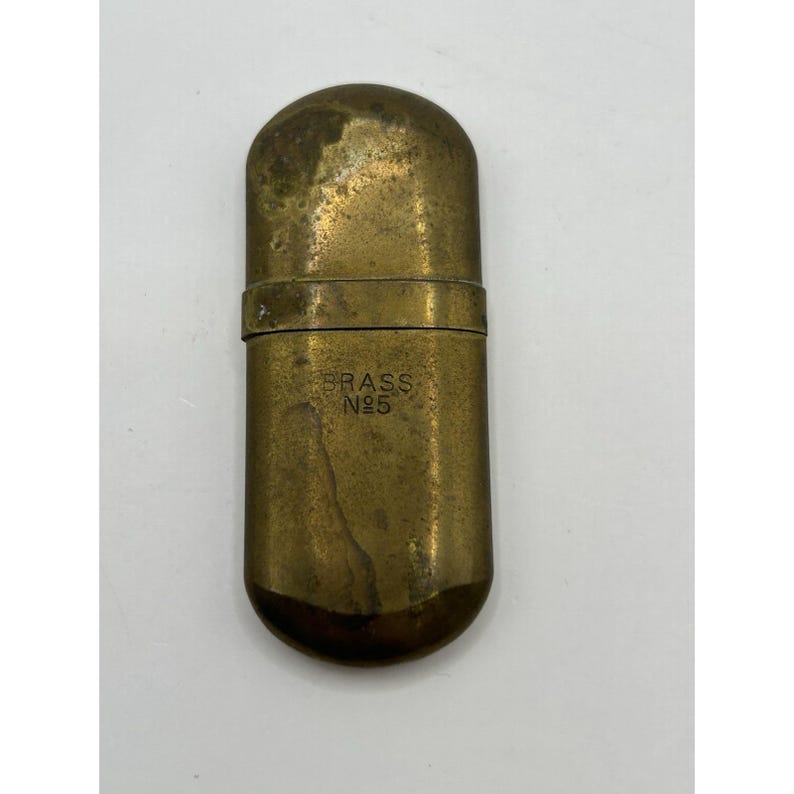 Puede incluir: Un estuche rectangular de lat&oacute;n con extremos redondeados y tapa con bisagra. La superficie tiene una p&aacute;tina desgastada y deslustrada. El frente del estuche est&aacute; grabado con el texto BRASS No5 en may&uacute;sculas, indicando el material y modelo.