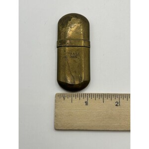 Puede incluir: Un estuche rectangular de lat&oacute;n con extremos redondeados y una p&aacute;tina desgastada. El frente est&aacute; estampado con el texto BRASS NO5. Una regla de madera se coloca horizontalmente debajo del objeto, mostrando que mide unos 5,7 cm de largo.