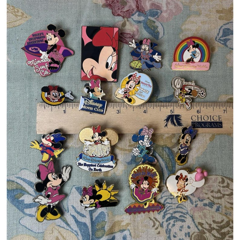 May include: A collection of colorful Minnie Mouse pins, featuring various designs and themes. Some pins include text like "Minnie Surf Co." and "Disney Movie Club." The pins are arranged on a patterned surface, with a ruler for scale.