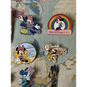 May include: A collection of Disney Minnie Mouse enamel pins. The pins feature Minnie Mouse in various outfits and settings, including a rainbow, a beach scene, and a scuba diving outfit. The pins are colorful and detailed.