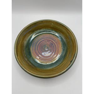 May include: A round ceramic bowl featuring a glazed finish with a blend of earthy brown and deep green tones. The center of the bowl displays a distinct circular spiral pattern with iridescent purple and copper hues radiating outward.