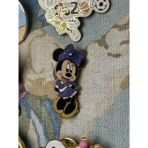 May include: A Minnie Mouse enamel pin in a purple dress and bow. The pin has a gold border and black shoes. Other pins are visible in the background.