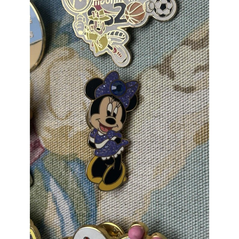 May include: A Minnie Mouse enamel pin in a purple dress and bow. The pin has a gold border and black shoes. Other pins are visible in the background.