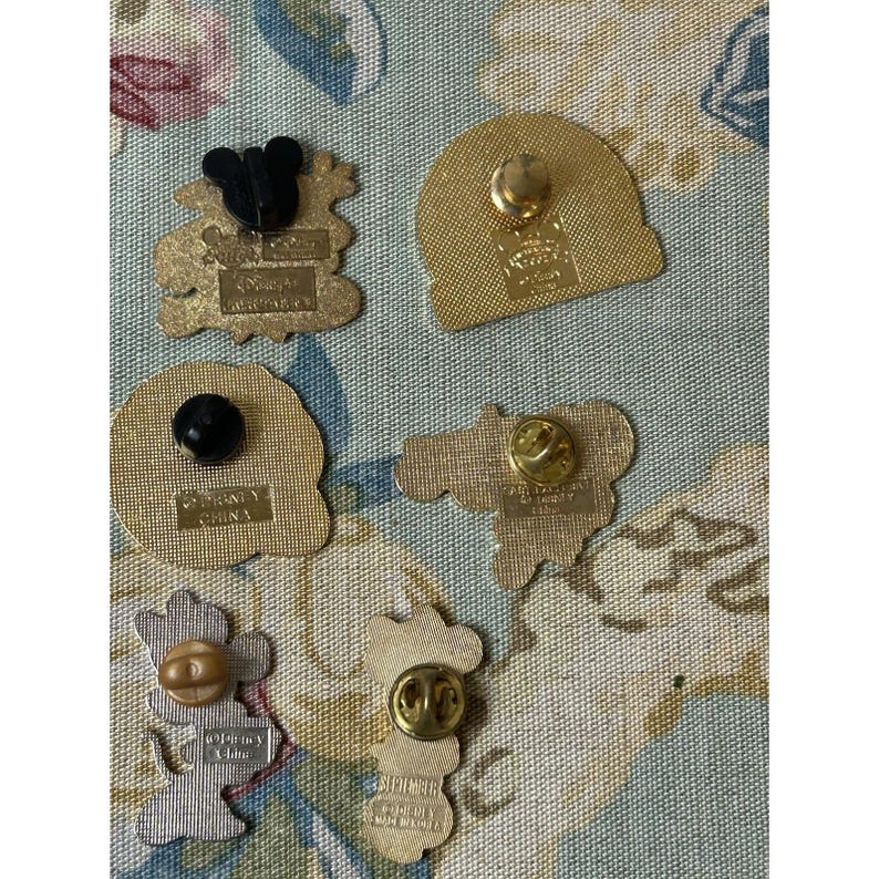May include: A collection of five Disney pins with various shapes and designs. The pins feature Mickey Mouse, Minnie Mouse, and other characters. The pins are made of metal and have a gold finish. The backs of the pins have a pin clasp.