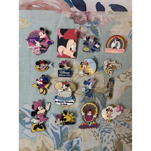 May include: A collection of Disney Minnie Mouse pins in various designs. The pins feature Minnie in different outfits and poses, with some including text like "Disney Movie Club" and "The Happiest Celebration On Earth". The pins are colorful and detailed.