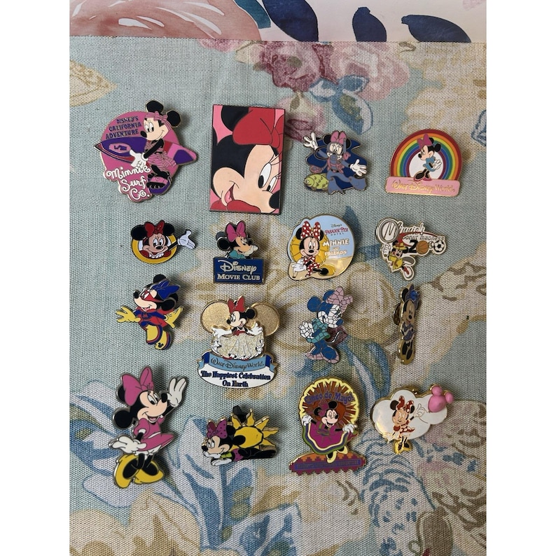 May include: A collection of Disney Minnie Mouse pins in various designs. The pins feature Minnie in different outfits and poses, with some including text like "Disney Movie Club" and "The Happiest Celebration On Earth". The pins are colorful and detailed.