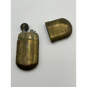 Puede incluir: Un encendedor de lat&oacute;n vintage con cuerpo rectangular y bordes redondeados. El encendedor se muestra con su tapa de lat&oacute;n a juego retirada, revelando la rueda de encendido interna y la mecha. La superficie met&aacute;lica muestra signos de desgaste.