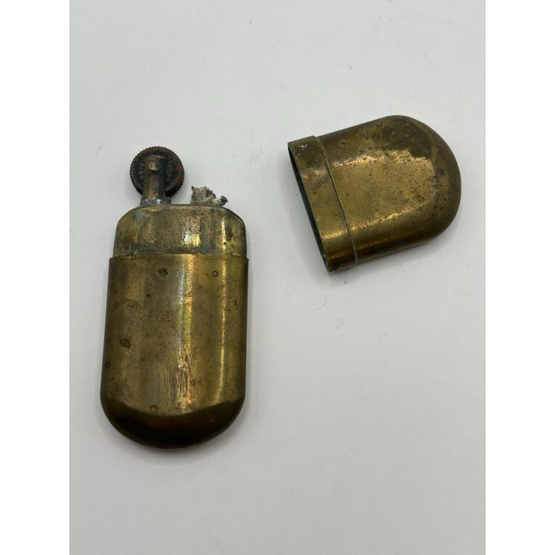 Puede incluir: Un encendedor de lat&oacute;n vintage con cuerpo rectangular y bordes redondeados. El encendedor se muestra con su tapa de lat&oacute;n a juego retirada, revelando la rueda de encendido interna y la mecha. La superficie met&aacute;lica muestra signos de desgaste.