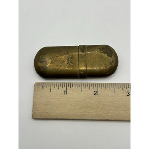 Puede incluir: Un estuche oblongo de lat&oacute;n con extremos redondeados y una banda central, que presenta el texto estampado BRASS N&deg;5. Una regla de madera se coloca directamente debajo del objeto, indicando una longitud de unos 7 cm para el recipiente.