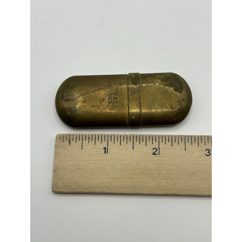 Puede incluir: Un estuche oblongo de lat&oacute;n con extremos redondeados y una banda central, que presenta el texto estampado BRASS N&deg;5. Una regla de madera se coloca directamente debajo del objeto, indicando una longitud de unos 7 cm para el recipiente.