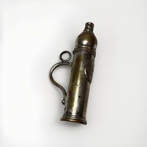 Può includere: Un flacone per polvere da sparo cilindrico in metallo vintage con finitura in ottone ossidato e toni argentati. L'oggetto presenta un collo stretto, un manico laterale curvo con un anello circolare attaccato e una superficie usurata e patinata.