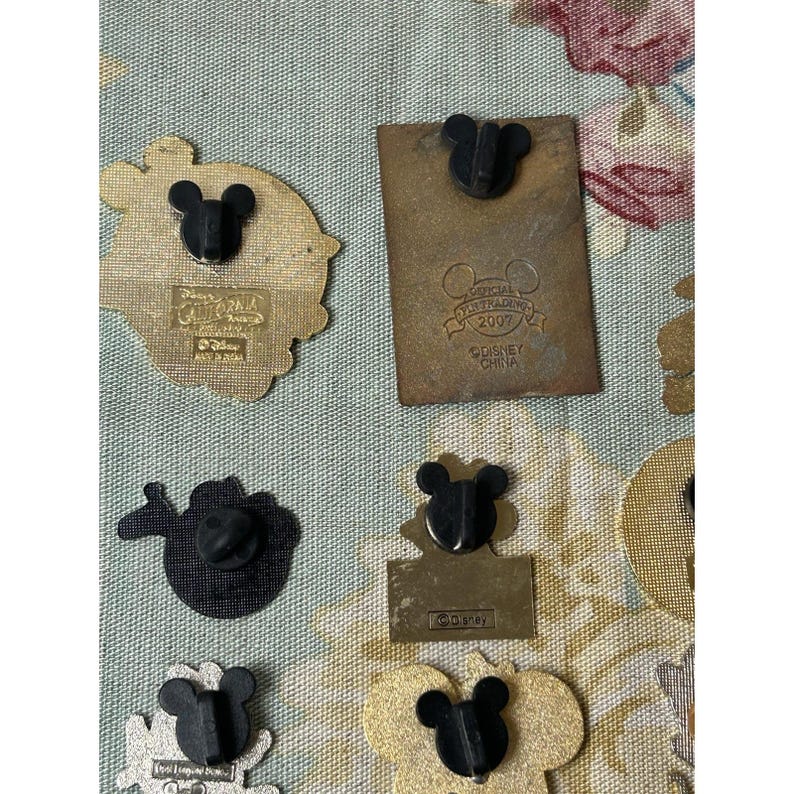 May include: A collection of Disney pins with black Mickey Mouse head designs. Some pins have gold and silver tones, while others are black. One pin reads "Official Pin Trading 2007 Disney China."