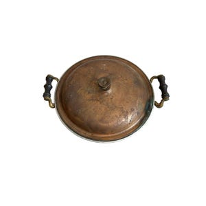 May include: A round copper pot with a lid, featuring a weathered, tarnished patina across the surface. The pot includes two side handles made of dark, turned wood attached with brass fittings and a small central knob on the lid.
