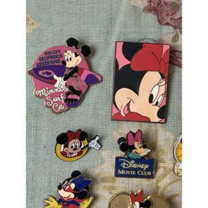 May include: Collection of Disney Minnie Mouse pins. One pin features Minnie Mouse surfing with the text "Disney's California Adventure" and "Minnie Surf Co." Another pin shows Minnie's face. Other pins include the Disney Movie Club logo.