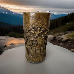 Könnte beinhalten: Ein zylindrisches, geschnitztes Holzgefäß mit einem detaillierten Relief, das einen Baum, Gebäude und Figuren darstellt. Das Gefäß hat einen warmen, goldbraunen Farbton und ist vor einem verschwommenen Bergpanorama platziert.