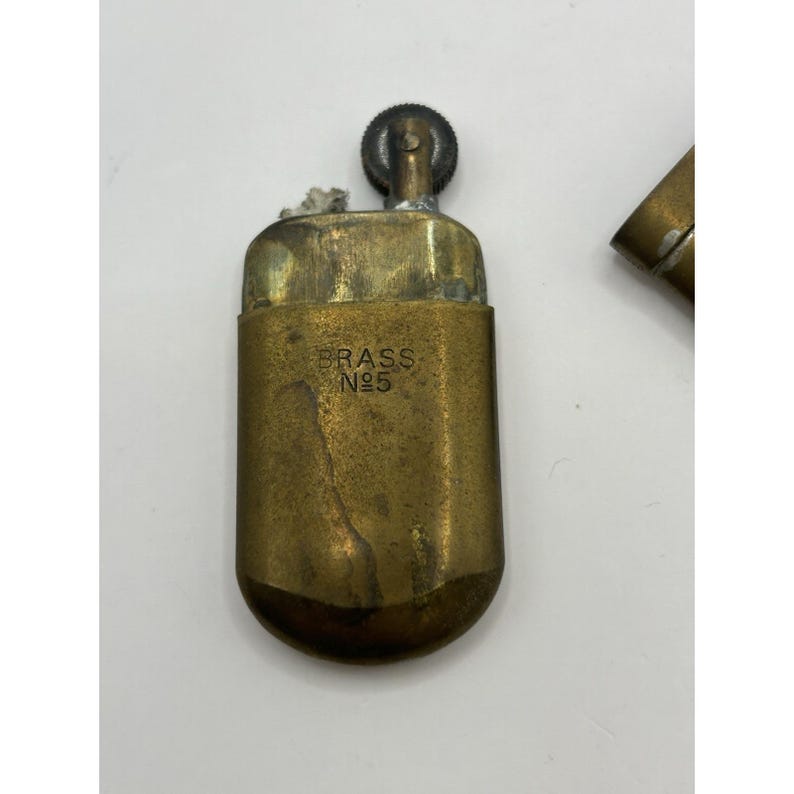 Puede incluir: Un encendedor rectangular de lat&oacute;n con esquinas inferiores redondeadas, que presenta una rueda de encendido texturizada en la parte superior y el texto grabado BRASS No5 en la parte frontal. El metal muestra signos de desgaste y oxidaci&oacute;n.