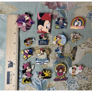 May include: A collection of Minnie Mouse pins in various designs. Some pins feature Minnie in different outfits, while others have text like "Disney Movie Club" and "California Adventure". The pins are colorful and detailed.