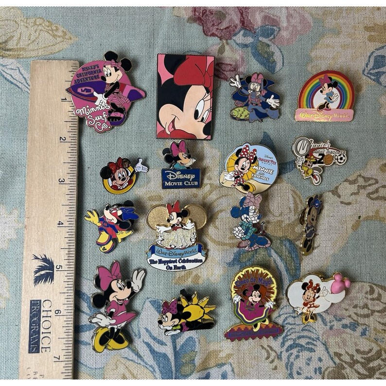 May include: A collection of Minnie Mouse pins in various designs. Some pins feature Minnie in different outfits, while others have text like "Disney Movie Club" and "California Adventure". The pins are colorful and detailed.