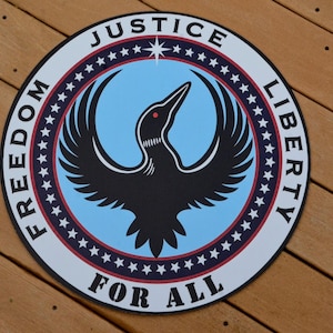 May include: A circular sign featuring a black loon silhouette with a red eye centered on a light blue background. A dark border contains the white text JUSTICE, LIBERTY, FREEDOM, FOR ALL and a ring of small white stars against a dark red background.