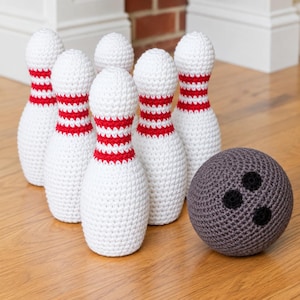 Può includere: Un set di sei birilli da bowling all'uncinetto bianchi con strisce rosse orizzontali attorno al collo e una singola palla da bowling all'uncinetto grigia con tre fori neri circolari disposti a triangolo sulla superficie.