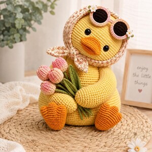 May include: A yellow crocheted duck toy wearing a floral headscarf and pink-rimmed sunglasses. The duck has orange feet and a beak, and it is holding a small bouquet of pink tulips with green stems. The toy features black bead eyes.