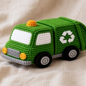 Pode incluir: Um caminhão de lixo de brinquedo feito em crochê verde, com janelas brancas, uma luz amarela no teto, para-choque cinza e rodas pretas com centros cinzas. A lateral do caminhão exibe um símbolo de reciclagem branco bordado no corpo.