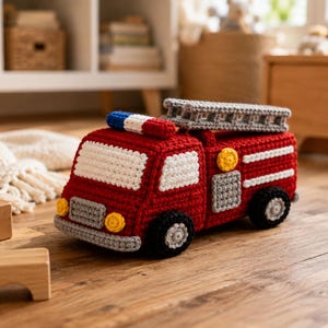 May include: A crocheted fire engine toy in vibrant red with white windows, a grey ladder on top, and a blue and white light bar. The vehicle features black and grey wheels, yellow circular headlights, and grey accents on the side and front bumper.