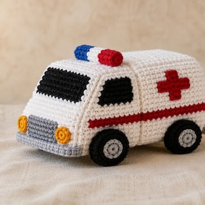 May include: A crocheted toy ambulance in white with a red cross on the side, a red horizontal stripe, black windows, and black wheels with grey centres. The vehicle features a red and blue light bar on the roof and yellow circular headlights.