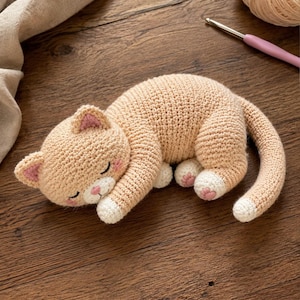 May include: A crocheted stuffed cat toy in a sleeping position, featuring light beige yarn, white paws, a white muzzle, pink inner ears, and pink cheek accents. The cat has closed eyes and a small pink nose, with a long tail curled to the side.