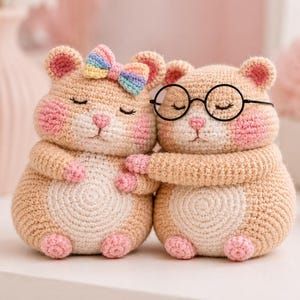 May include: Two crocheted hamster plushies in a beige yarn with pink accents on the cheeks, paws, and feet. One hamster wears a small rainbow-colored bow on its head, while the other wears round black-rimmed glasses. Both have closed eyes.