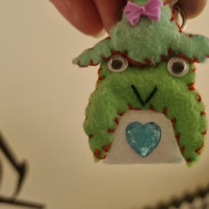May include: A handmade felt keychain shaped like a green creature with a light green hat, a small purple bow, googly eyes, and a stitched V-shaped mouth. The body features a white belly patch with a central blue heart-shaped sequin accent.