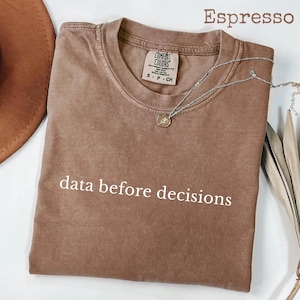 May include: A brown short-sleeve t-shirt with the text "data before decisions" printed in white on the front. The shirt features a crew neckline and a Comfort Colors brand label inside the collar. A silver chain necklace rests on the fabric.