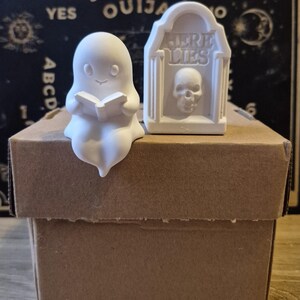 May include: A white matte figurine of a ghost reading a book and a white matte tombstone figurine with a skull relief and the text HERE LIES, both positioned on top of a brown cardboard box. The ghost has a smooth surface and simple facial features.